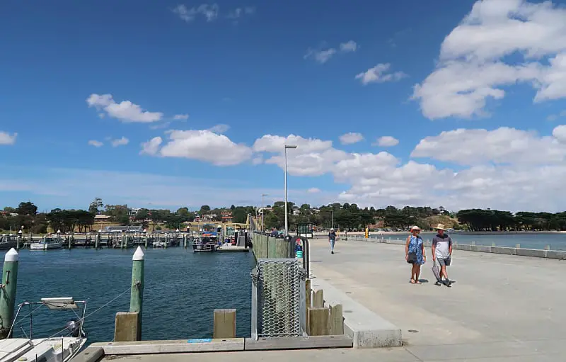 Visitors stroll along the spacious Portarlington Pier, with the marina and moored boats in the background, under a blue sky dotted with fluffy clouds.