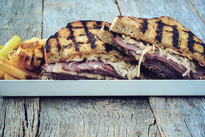 A toasted Reuben Sandwith filled with corned beef, swiss cheese, and sauerkraut sitting on a timber table.