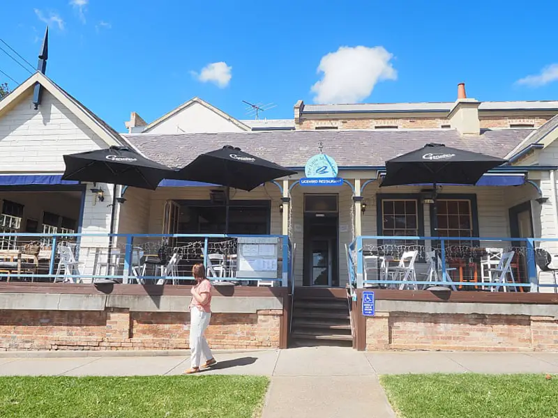 Exterior view of The Little Mussel Café on a sunny day, featuring a quaint building with a blue sign, outdoor seating under black umbrellas, and a clear blue sky above. A person is walking by the front entrance.