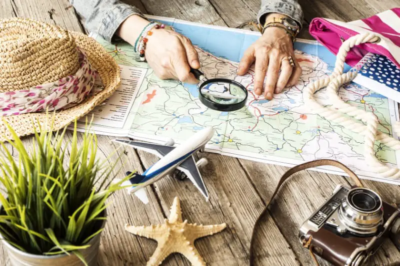 View of a person's hands holding a magnifying glass over a map. There is a model plane and starfish sitting on the table. Image of a person planning their  Victoria trip.