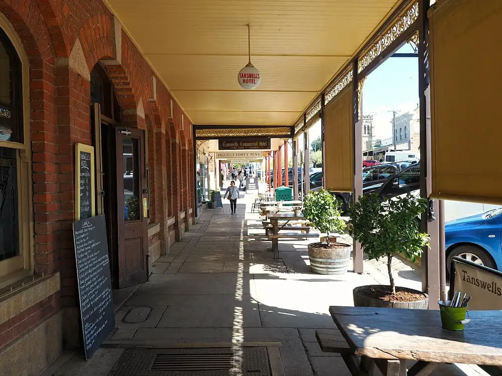 Footpath with a verandah at Beechworth in Victoria Australia. There are large potted plants, a table and chairs, and a woman walking along the street.