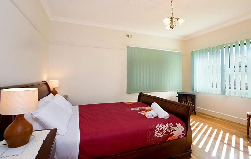 Bedroom at Bella Vista. There is a double bed made up with a red blanket and two windows with green vertical blinds.