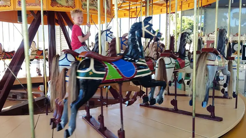 Young boy riding the carousel at the Geelong Waterfront. He is sitting on a white horse next to a black horse with a green and blue saddle.