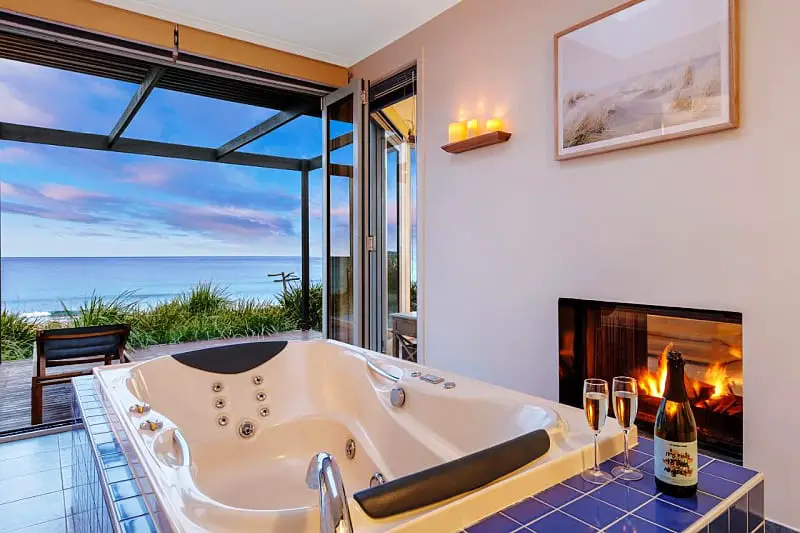 Bathroom with a spa bath, gas fireplace, and open glass doors with ocean views at Chocolate Gannets. There is a bottle of champagne and champagne glasses beside the bath