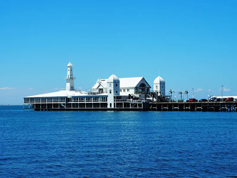 View of the restaurants at Cunningham Pier. The buildings are bright white against a blue sky and bay. Palm trees can be seen being blown by the wind.