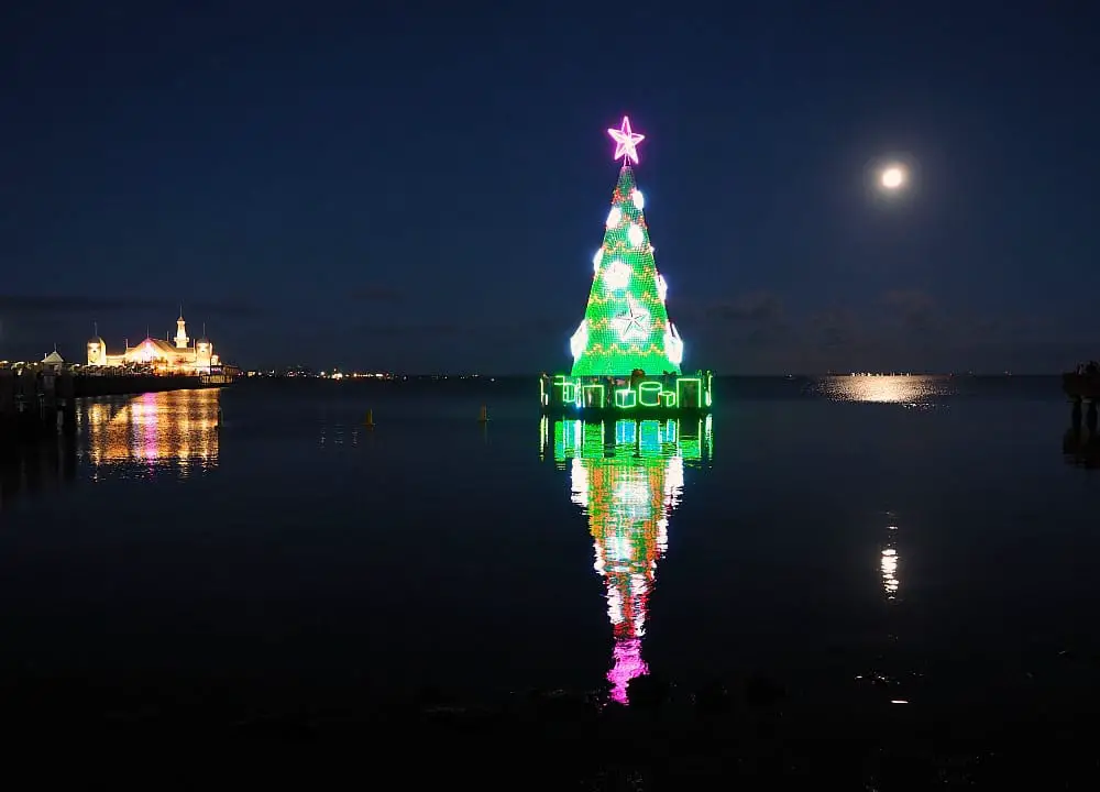 The Geelong Floating Christmas Tree at night with green lights and a pink star. The lights of Cunningham Pier can be seen in the background. 