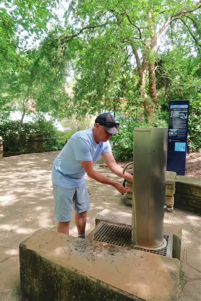 Man testing the water at Hepburn Mineral Springs. The water fountain is surrounded by shady trees. Visiting the mineral springs is one of the top things to do in Daylesford.