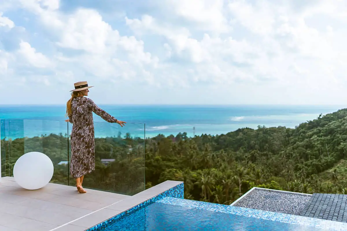 Woman in a long dress and hat standing on a deck overlooking the coastal forest with panoramic ocean views at luxury accommodation on the Great Ocean Road.
