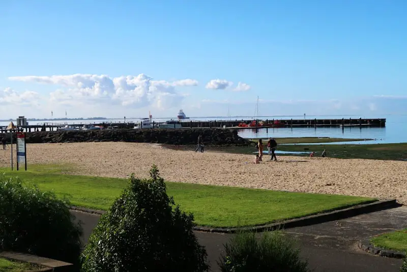 People enjoying themselves at Rippleside Beach on the western edge of Geelong Waterfront. A breakwater can be seen and there is a boat out in the bay. There is a small patch of lush grass in front of the beach.