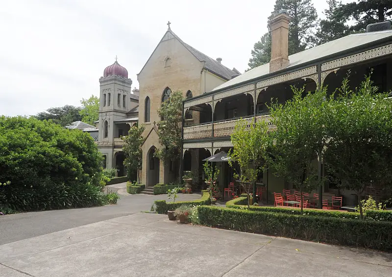 View of The Convent Art Gallery and Cafe in Daylesford. There are 3 old buildings including an old convent and a historic two storey house with verandahs  and decorative fretwork. There is a driveway and garden in the foreground. 