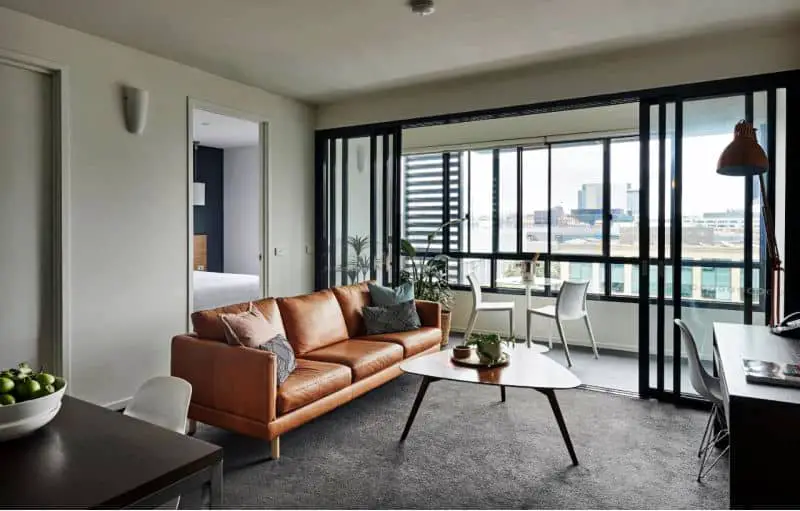 Guest room at the Vue Apartments at Geelong Waterfront with an enclosed balcony. There is a tan leather couch and oblong coffee table in the living area.
