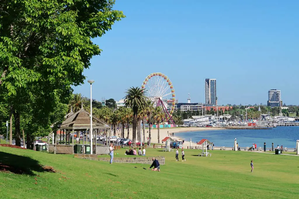 View of people relaxing on the sloping lawns at Waterfront Geelong. Geelong Beach, the city skyline, and the Giant Sky Wheel can be seen in the distance.
