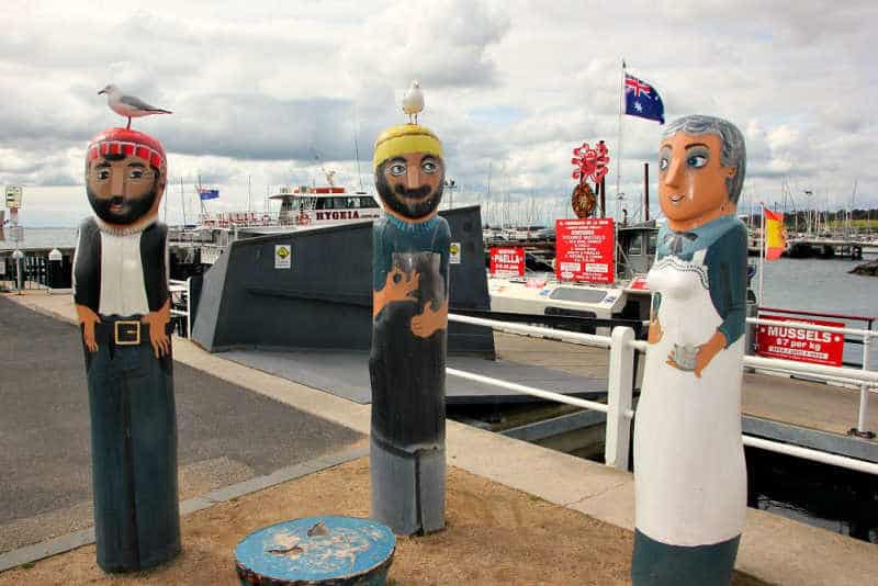 Three characterful bollards painted to look like people - a sailor, a fisherman, and a woman in a dress and apron - stand by a harbour with boats and an Australian flag in the background, under a cloudy sky. Seagulls perch on the heads of the sailor and fisherman.