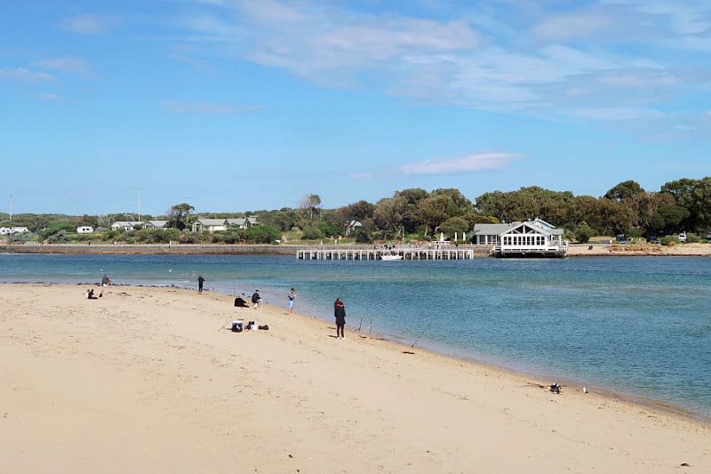A serene beach scene along the Barwon Heads River Beach, with individuals and small groups scattered across the sand, some fishing. A classic white beach house with a long pier extends over the calm blue water, set against a backdrop of clear skies and lush greenery.
