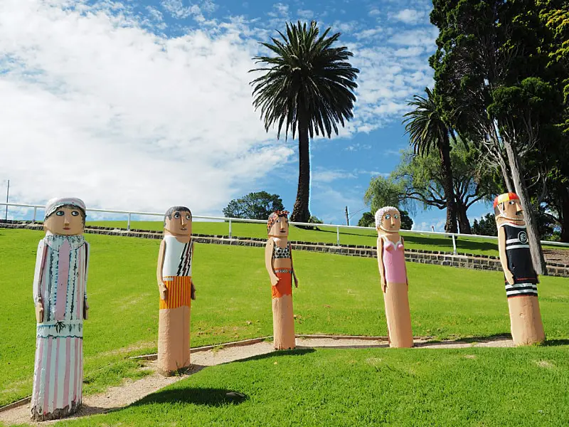 A row of colorful, stylized sculptures resembling people in an outdoor setting, with a tall palm tree and lush greenery in the background, under a bright blue sky.