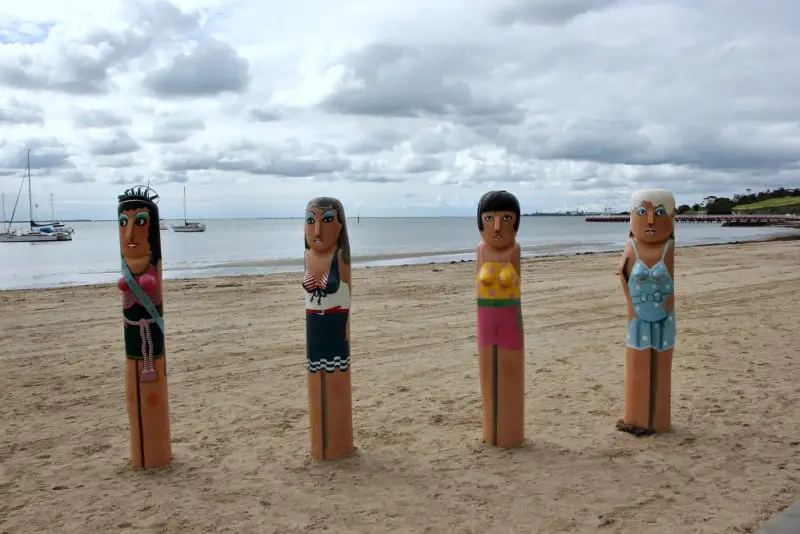 Four painted Geelong bollard figures, each with unique swimwear designs, stand on the sandy Eastern Beach with moored sailboats in the calm sea and a cloudy sky in the distance.
