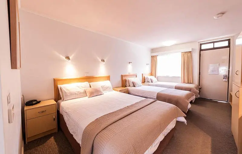 A cozy and inviting guest room at Eastern Sands Geelong Waterfront motel accommodation featuring a large double bed with beige bedding and a single bed by the window, both with wooden headboards and soft lighting, offering a comfortable stay in a well-lit, neutral-toned space.
