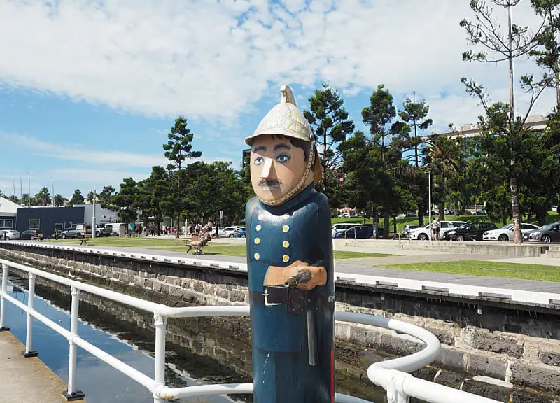 A bollard sculpture representing an 1800s fireman in traditional service attire stands holding a hose beside a waterway, with a white railing, parked cars, pedestrians, and tall green trees under a blue sky with scattered clouds in the background.