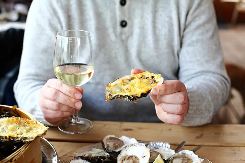A patron at Fishermens Pier restaurant on the Geelong waterfront is depicted in a casual grey sweater, savoring the local cuisine with a grilled oyster in the left hand prominently shown to the camera. The right hand holds a glass of white wine, complementing the array of fresh oysters garnished with lemon wedges on the wooden table. Fishermens Pier is one of the best Geelong waterfront restaurants for lovers of seafood.