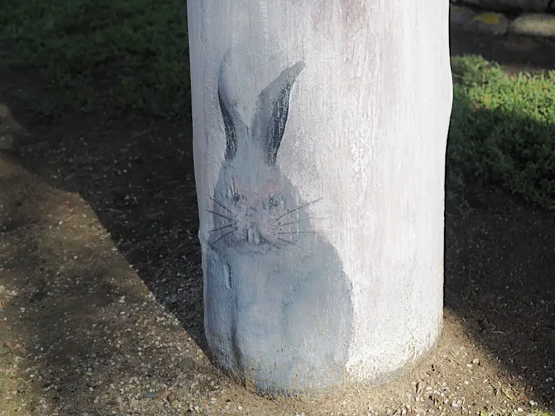 A painting of a blue rabbit on a cylindrical bollard post, with subtle shadows cast on the ground indicating sunlight from the upper left.