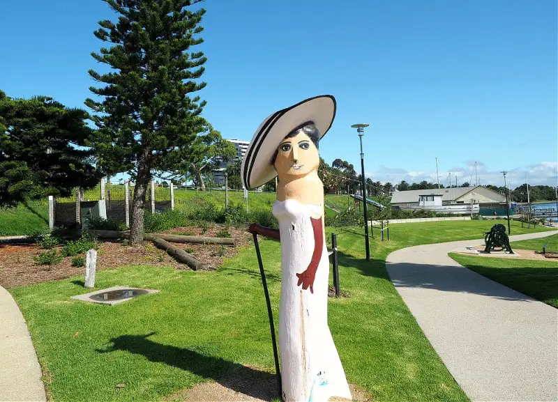 A painted Geelong Bollard resembling a woman in a white dress with a wide-brimmed hat and red gloves stands beside a winding path in a sunny park, with pine trees and a glimpse of a harbor in the distance. The playful art installation adds character to the tranquil outdoor setting.