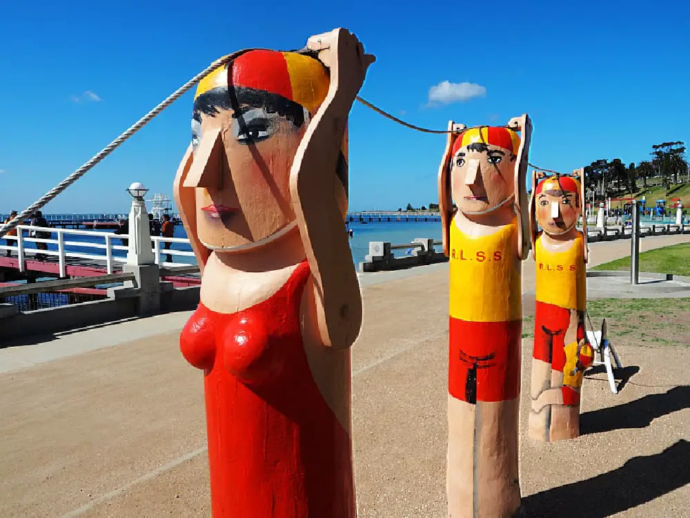 The colourful Geelong Bollards sculptures depicting lifesavers in red and yellow with the acronym 'R.L.S.S' painted on their torsos line the sunny Geelong Baywalk with a clear blue sky overhead and the Eastern Beach Promenade extending into the sea in the background.