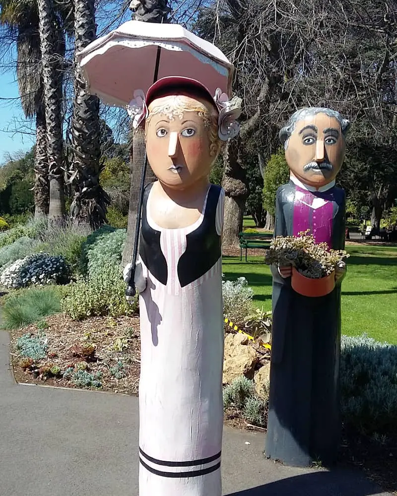 Two whimsical Geelong Bollards in a garden setting with the female foreground figure sporting a frilled parasol, a pink striped dress, and a bonnet, and the male background figure in a purple and black suit holding a flower pot.