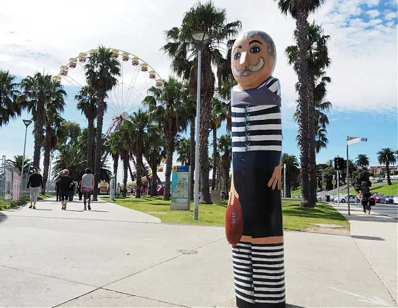 A Geelong Baywalk Bollard sculpture of an early Australian Rules Football Geelong player. A man in blue and white horizontal striped outfit with a football in his hand stands along a palm-lined pathway near a Ferris wheel under a clear blue sky.