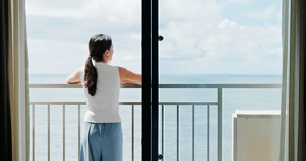 A woman stands on a balcony at Geelong Waterfront accommodation, overlooking a calm Corio Bay under a clear blue sky.