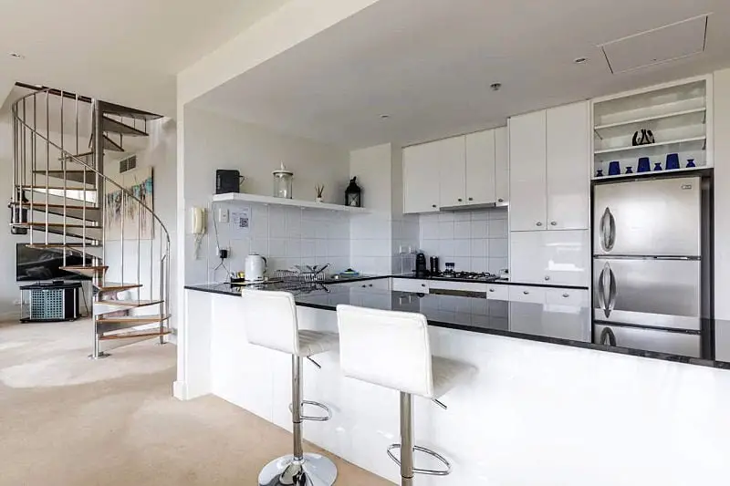 Modern kitchen in a Geelong Waterfront Penthouse Apartment featuring white cabinetry, stainless steel appliances, a gas cooktop, and a spiral staircase leading to an upper level.