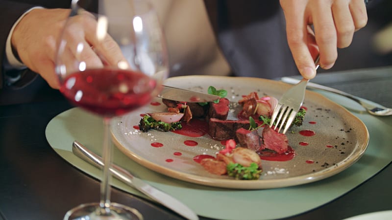 Close-up of a diner enjoying a gourmet meal at La Cachette, with a focus on a plate of beautifully presented sliced meat, garnished with green herbs and red sauce dots. A hand is seen holding a fork and a glass of red wine, suggesting an elegant dining experience.