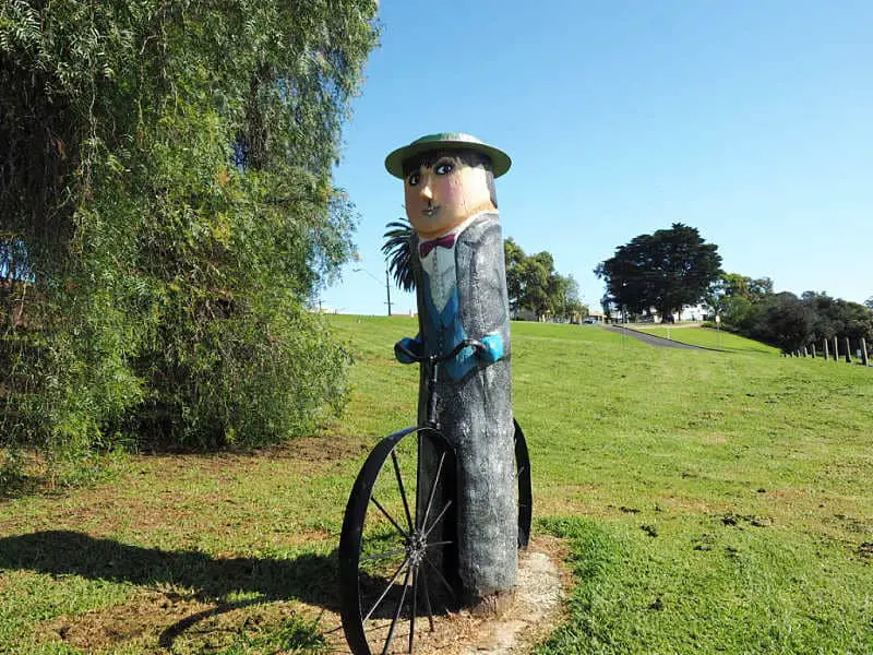 A whimsical  Geelong bollard sculpture dressed in a vintage suit and bow tie, holding a bicycle wheel, stands in a green park under a clear sky, with lush trees and a gently sloping hill in the background.