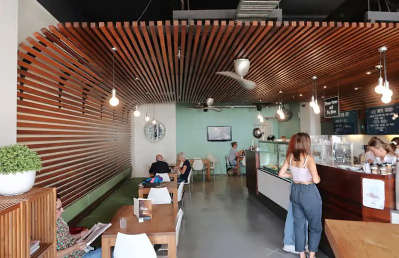 Interior of the modern Panache cafe with customers seated at tables, featuring a distinctive wooden slat design on the ceiling and a counter with a display case. The ambient lighting and mint green walls create a relaxed atmosphere.