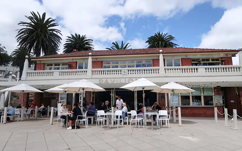 Outdoor seating area at Pavilion Geelong Waterfront cafe with patrons dining under white umbrellas, set against a backdrop of palm trees and a clear sky, with the cafe's two-storey building featuring a balcony in the background.