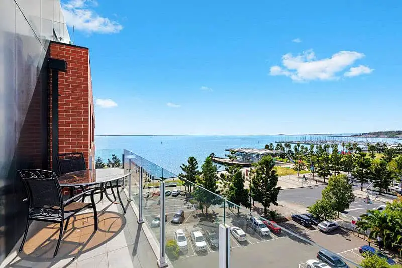 View from the Pierpoint 105 Waterfront Geelong apartment balcony featuring a patio set, with a panoramic vista of the ocean, a bustling street with parked cars, palm trees, and a pier extending into the tranquil blue waters of Corio Bay under a sunny sky.