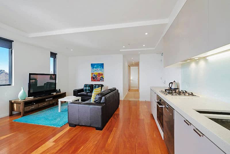 Interior of the Pierpoint 105 Waterfront Geelong apartment showing a modern, open-plan living space with polished wooden floors, a sleek kitchen counter, contemporary furniture including a dark grey sofa, and a vibrant painting adding a pop of color.