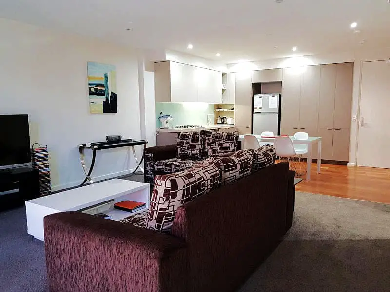 Interior of Pier Point 401 Waterfront Geelong holiday apartment featuring a contemporary open-plan living area with a brown textured sofa, modern white dining chairs, a kitchen in the background, warm wooden flooring, and soft ambient lighting.