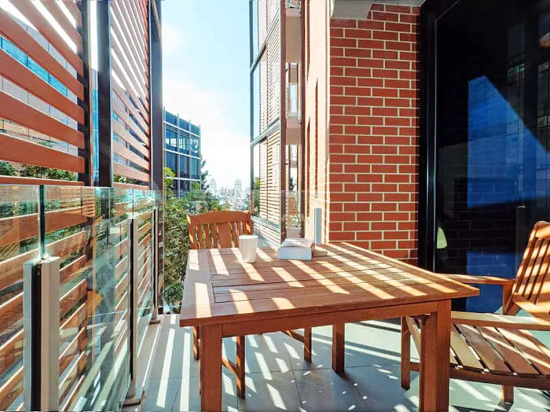 A sunny balcony setting at Pier Point 401 in Geelong with a wooden table and chairs, framed by clear glass balustrades and red brick walls, overlooking urban architecture.