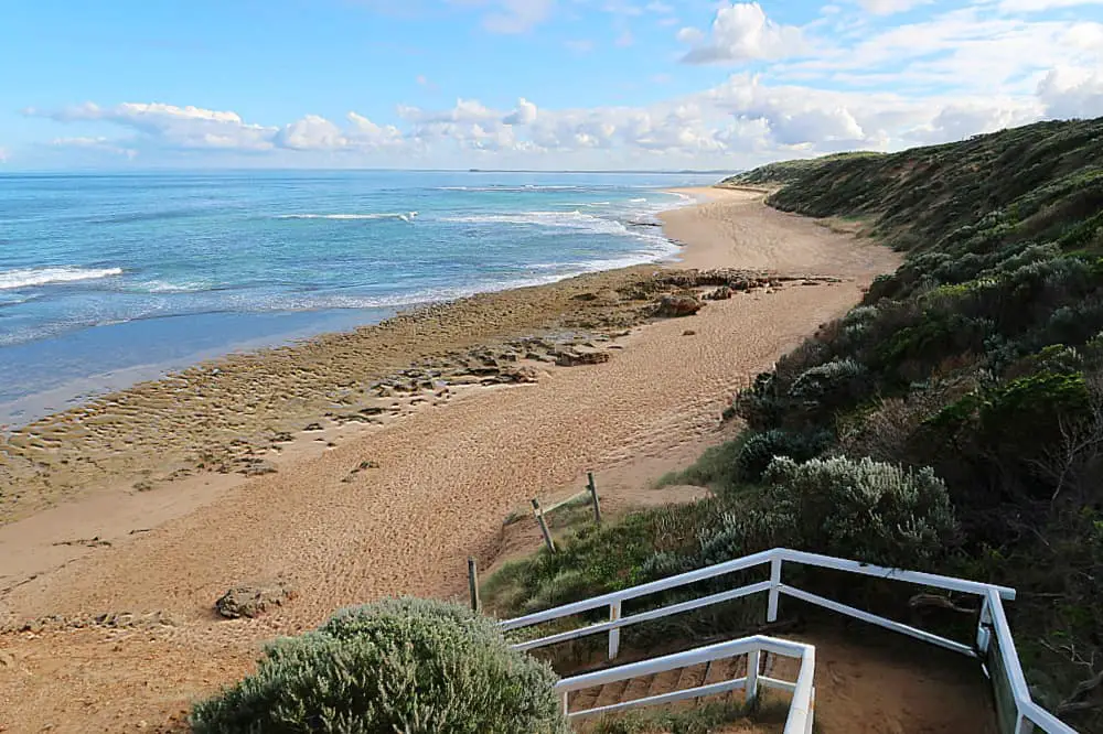 Elevated view of Point Lonsdale Beach, showcasing the coastline with rocky outcrops and sandy shores. The beach stretches into the distance, bordered by undulating dunes and native shrubbery, with a white wooden staircase leading down to the tranquil blue waters under a partly cloudy sky.
