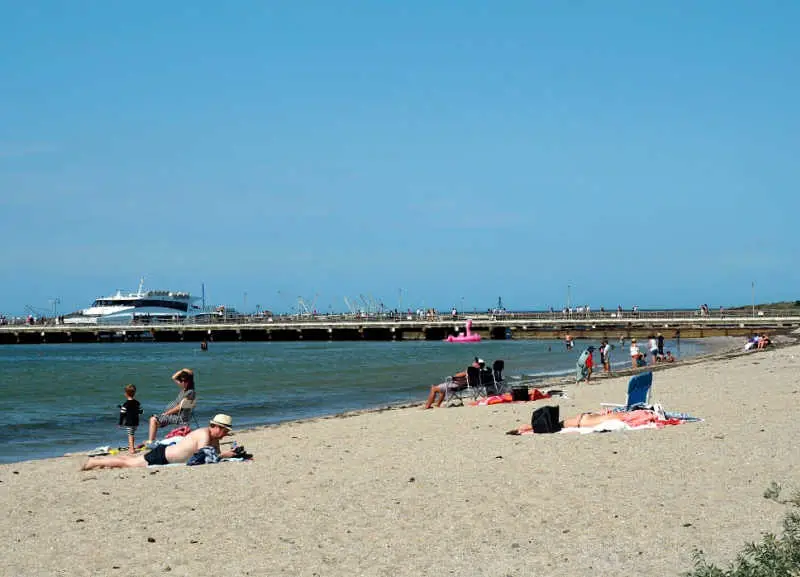 Busy summer day at Portarlington Beach with visitors relaxing on the sand and wading into the water. In the background, a Port Phillip Ferry is docked at a pier extending into the sea, under a clear blue sky, contributing to the lively coastal ambiance.
