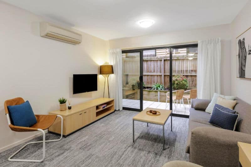 Cozy and well-lit living room interior at Quest Geelong Waterfront accommodation with modern furnishings, including a grey sofa, wooden TV stand, and floor lamp, leading to an outdoor patio area through glass sliding doors.
