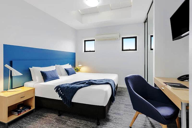 Quest Geelong modern bedroom featuring a king-sized bed with a bright blue headboard, crisp white linens, and coordinating throw pillows and blanket, complemented by a sleek wooden nightstand and a comfortable blue chair, all under the soft glow of ambient lighting.