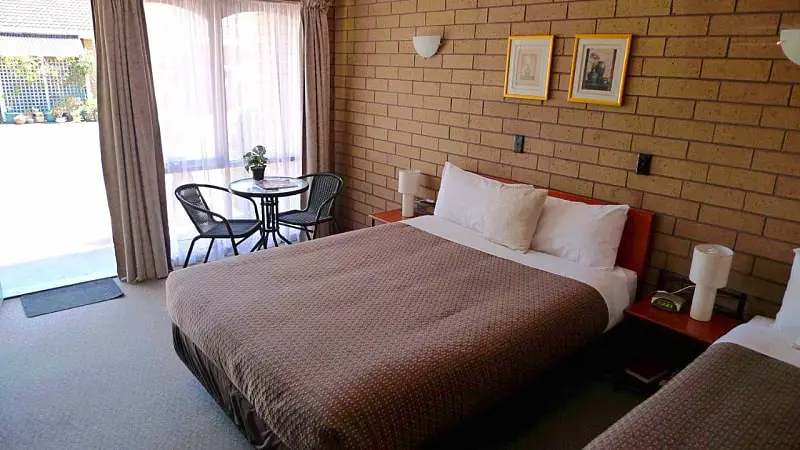 Inviting motel room at Rippleside Park featuring a large bed with a brown patterned bedspread, simple artwork on the brick wall, and a quaint dining area by the window with natural light streaming in.