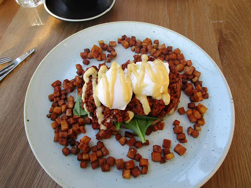 Picture of the Jack Sparrow breakfast at Sailors' Rest one of the best Geelong Waterfront cafes. Poached eggs drizzled with holandaise sauce on a bed of seasoned minced meat and spinach, surrounded by diced roasted potatoes, served on a ceramic plate with a casual dining setting in the background.