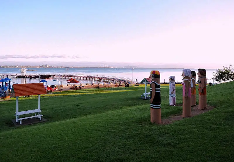 A peaceful Eastern Beach at sunrise with a row of colourful Geelong Bollards on a hill overlooking the calm Corio Bay with the promenade, playground, and distant city skyline.