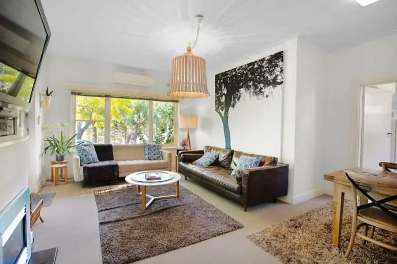 Bright and airy living room in the Beach House Apartment in Geelong, with a plush leather sofa, chic rattan chandelier, a wall mural of a tree, and a window nook offering a view of greenery outside.