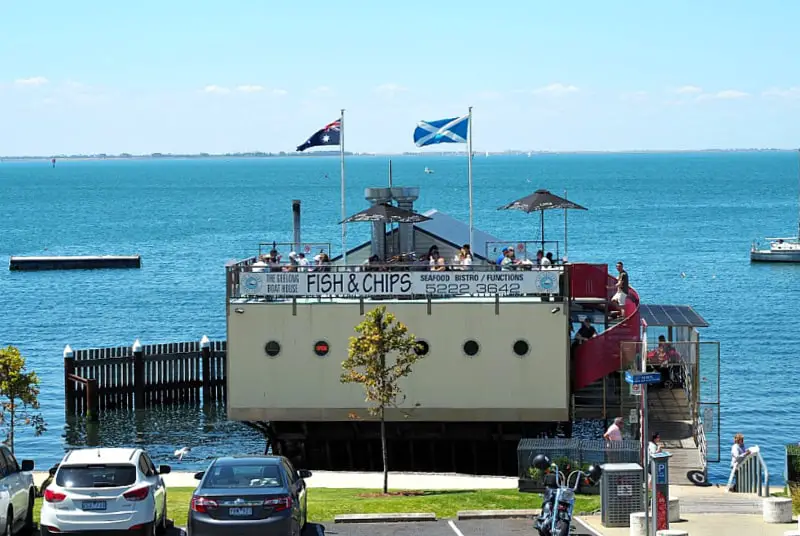 The Geelong Boat House, prominently advertised for its fish and chips, sits over the water, offering diners a picturesque view of the calm blue sea. Patrons can be seen enjoying their meals under umbrellas on the rooftop deck, while Australian and Scottish flags flutter in the breeze, enhancing the coastal vibe of this popular dining spot.