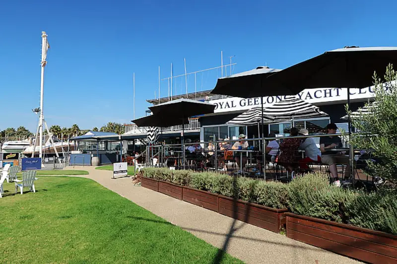 The Yot cafe at the Royal Geelong Yacht Club with diners seated on a raised deck under black and white striped umbrellas, beside a neatly manicured lawn with a white deckchair against a bright blue sky. 