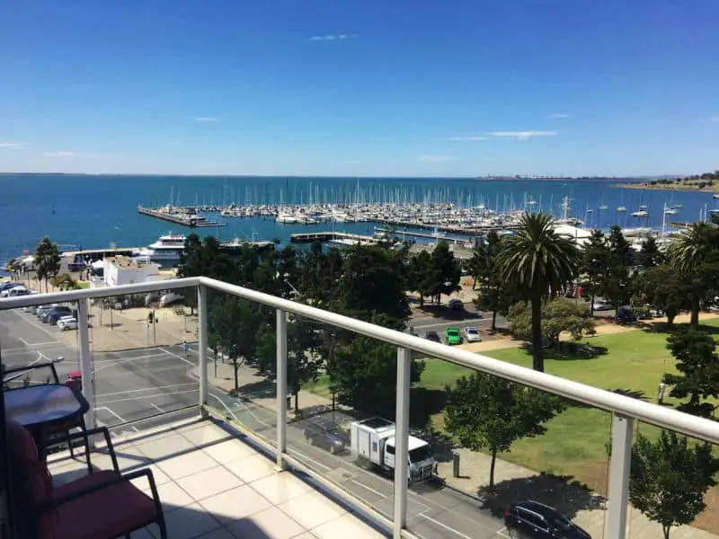 Balcony view from a Geelong Waterfront Penthouse Apartment, showcasing a bustling marina with numerous boats, lush green parkland, and a picturesque blue sky.
