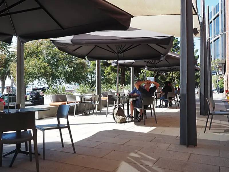 Outdoor patio area at the Waterfront Pantry cafe in Geelong with customers seated under large umbrellas on a sunny day surrounded by greenery there's a casual dining atmosphere.